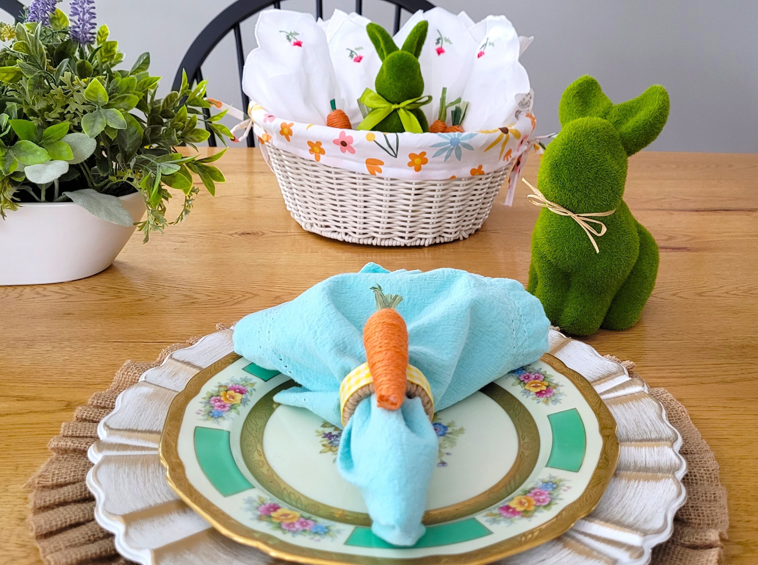 Easter place setting with white charger and floral plate with an aqua napkin in a jute carrot napkin ring A white basket with a green bunny and white napkins is in the background with another green bunny to the right side of the place setting