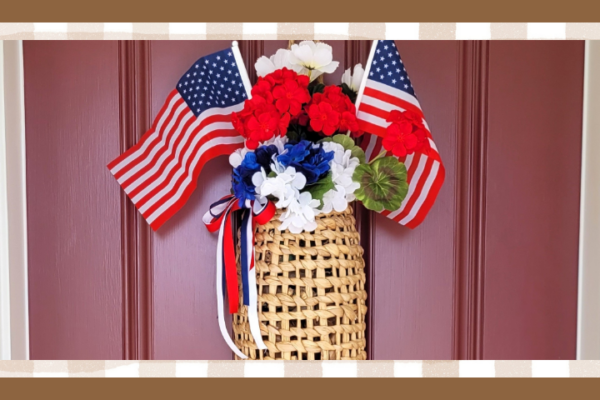 Basket filled with white and blue hydrangea and red geraniums Two small american flags are tucked in and red white and blue ribbons are tied on the side It hangs on a red front door