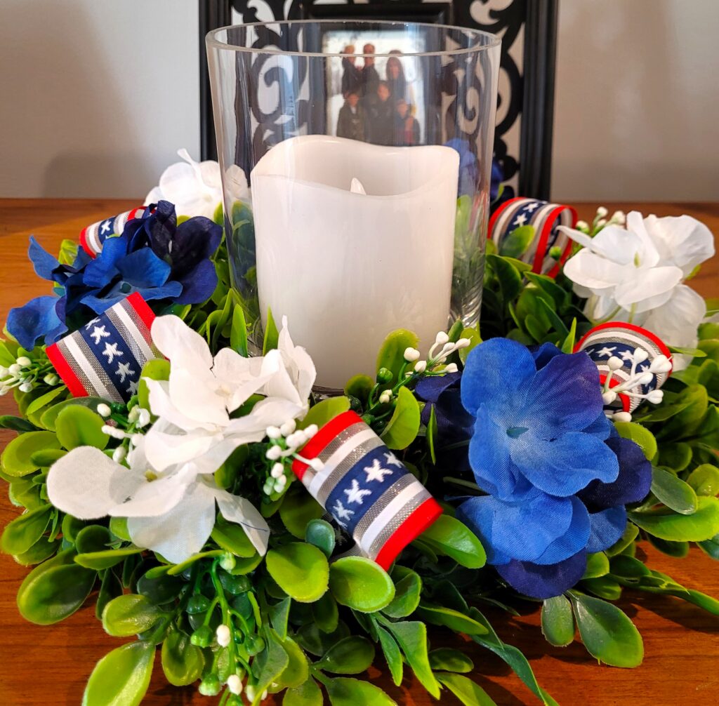 Memorial-Day-Candle-Ring | The Granite Acorn Boxwood candle ring with red white and blue ribbon tucked in along with blue and white hydrangea blooms A white pillar candle in a glass holder sits in the center