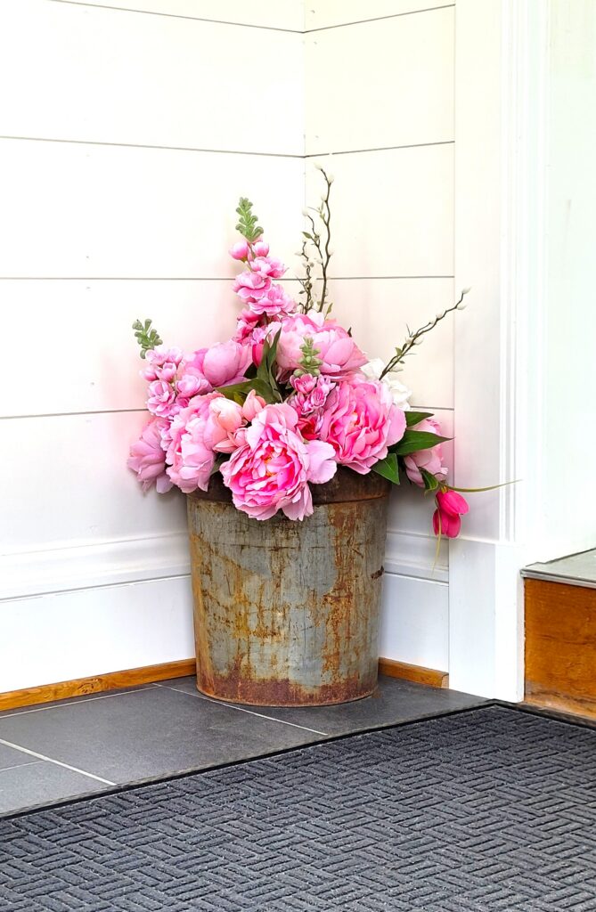 Rusty sap bucket filled with pink peonies tulips and snapdragons It's on a gray tiled floor against white painted shiplap.