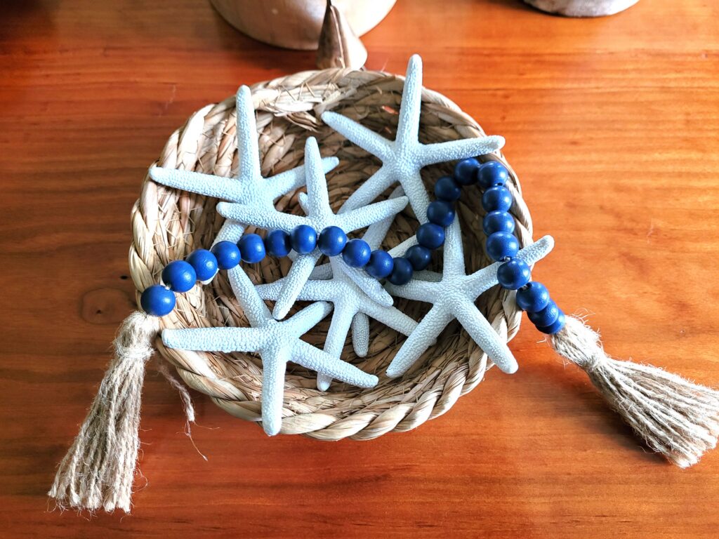 Starfish-Basket | The Granite Acorn Decorative white starfish pieces and a string of blue beads laid across a woven basket on a wooden surface