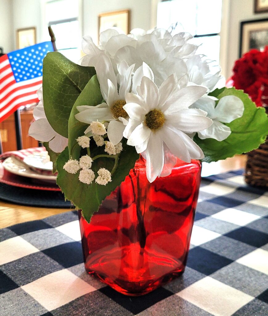 July-4-Flowers-Accent-2 | The Granite Acorn Red glass vase holds a bouquet of white daisies with greenery on a checkered table american flag in the background