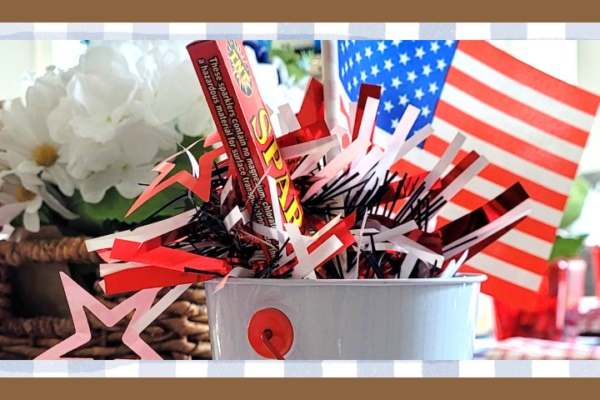 White bucket filled with red white and blue paper decorations and a small american flag with a floral arrangement and woven basket in the background