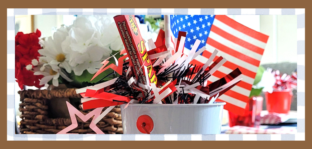 White bucket filled with red, white, and blue paper decorations and a small American flag, with a floral arrangement and woven basket in the background.