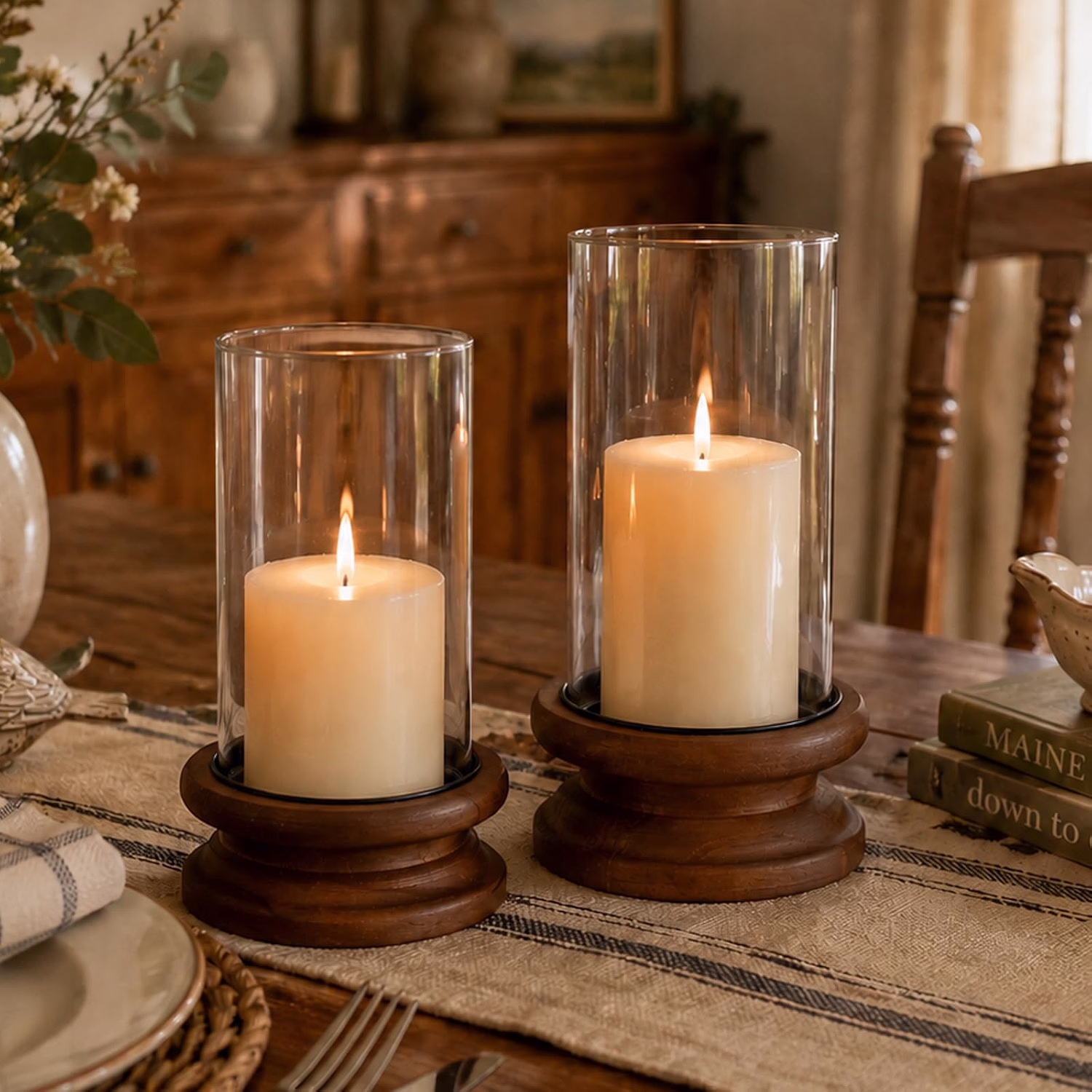 Two lit pillar candles inside glass hurricane holders sit on a rustic wooden dining table creating a warm centerpiece